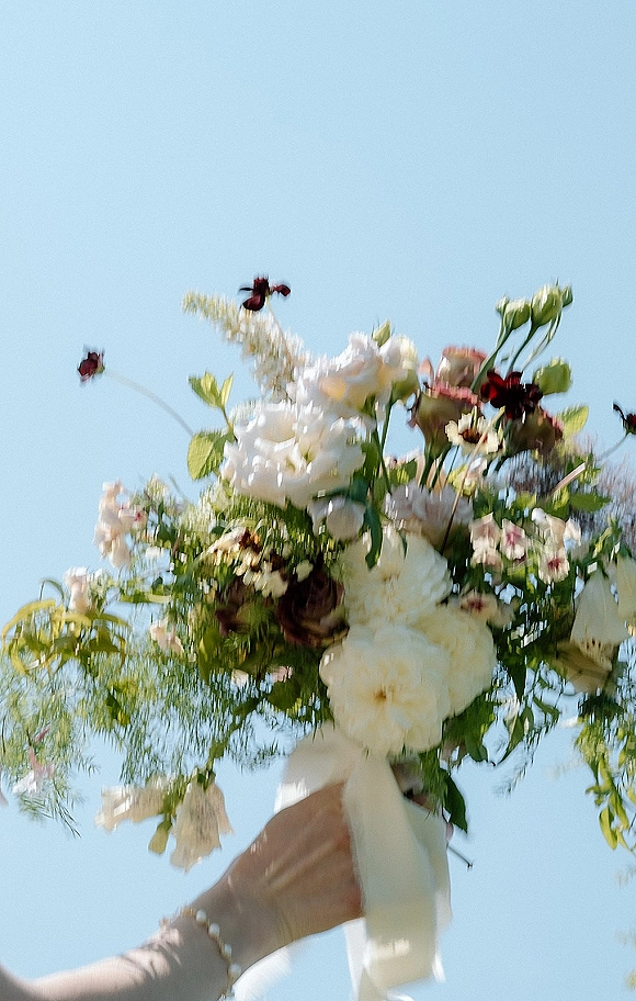 Bridal bouquet of white roses and mixed flowers with greenery and ribbon, held up against a blue sky with a pearl bracelet visible
