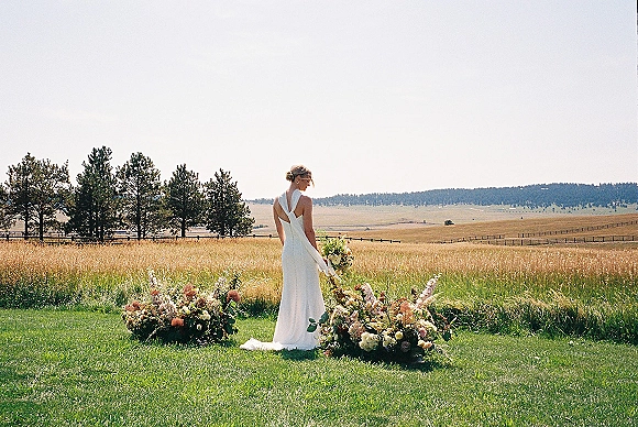 Bridal portrait of a bride from behind in a backless wedding dress holding a white-and-green bouquet in a wildflower meadow with fence