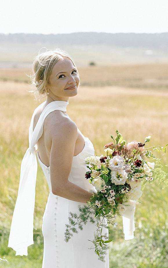 Bridal portrait of a smiling bride holding bouquet in an open back gown, looking over her shoulder in a tall grass field with hills