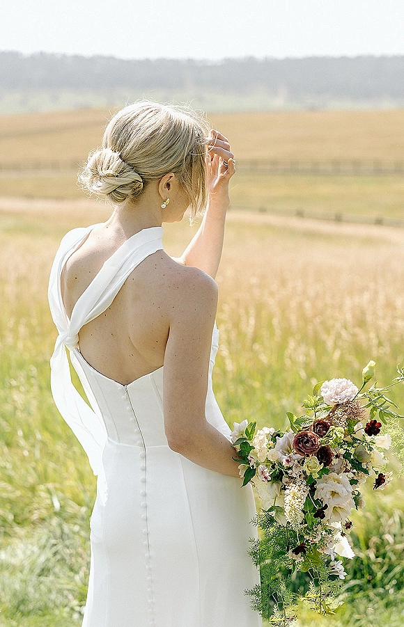 Bridal portrait of a bride from behind in a backless halter wedding dress, holding a wildflower bouquet in a tall grass field near farmland
