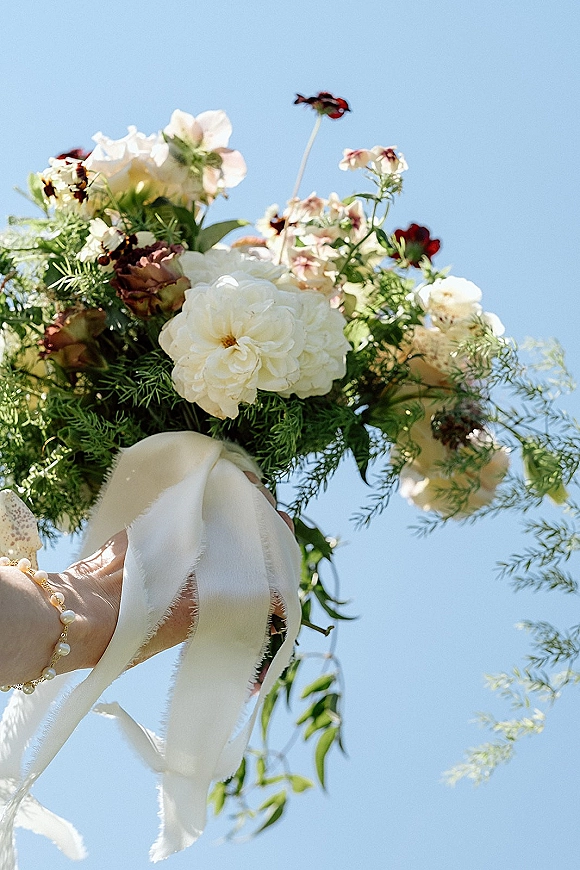 Bridal bouquet with white ribbon trailing from cream, blush, and burgundy blooms with greenery, held up in sunlight against blue sky