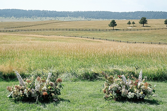 Ceremony altar flowers in low ground floral arrangements of white blooms, greenery, and dried florals set in a meadow with rolling hills