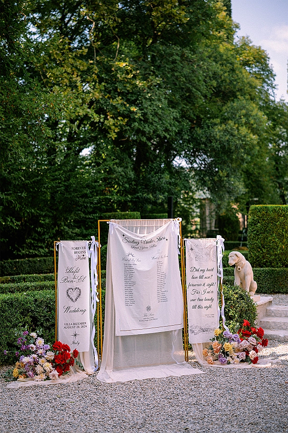 Wedding seating chart fabric banners with calligraphy text in gold frames, tied with ribbons and florals on a garden gravel path near stone steps