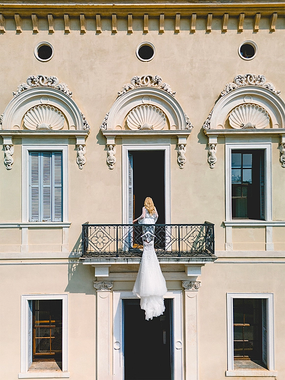 Bridal gown portrait of a bride in a long sleeve wedding dress on balcony, back view showing train, framed by ornate arched windows