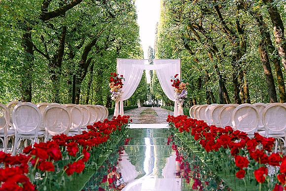 Outdoor ceremony setup with a white draped wedding arch and mirrored aisle runner lined with red roses along a tree-lined garden path