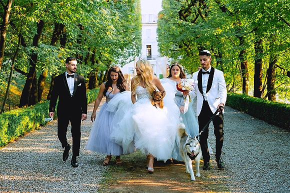 Wedding party portrait with bride and bridesmaids walking beside groomsmen on a tree-lined garden path, bride holding bouquet and dog leash