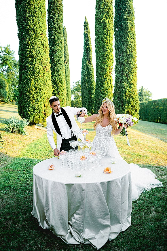 Champagne tower moment as bride in tiara and groom in white tux pour bubbly into coupe tower on lawn with cypress trees behind