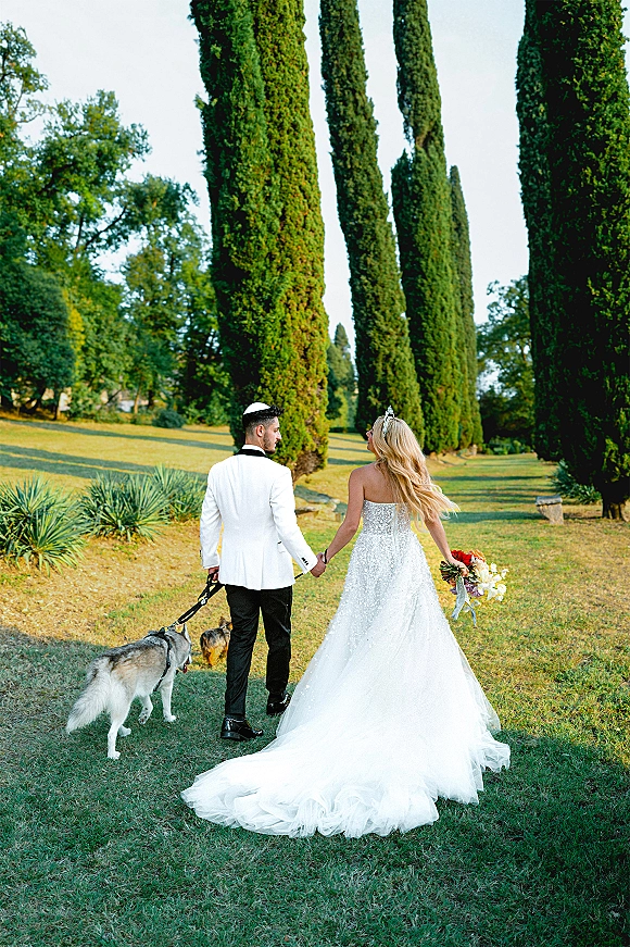Couple portrait of bride and groom walking away hand in hand, bride’s long train and bouquet, walking their dog in a cypress garden