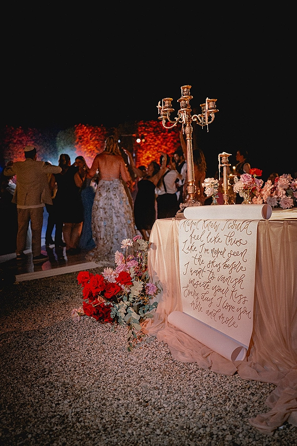 Wedding reception decor featuring a wedding welcome sign scroll with calligraphy, gold candelabra and candlelit florals as guests dance outdoors at night