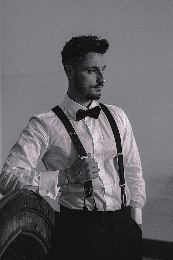 Groom portrait in black and white, groom with suspenders wearing a bow tie and cufflinks, leaning by a chair against a plain wall indoors