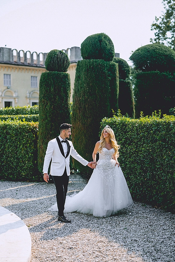 Couple portrait of bride and groom holding hands, bride in a tiara and strapless gown beside groom in tuxedo on a formal garden path