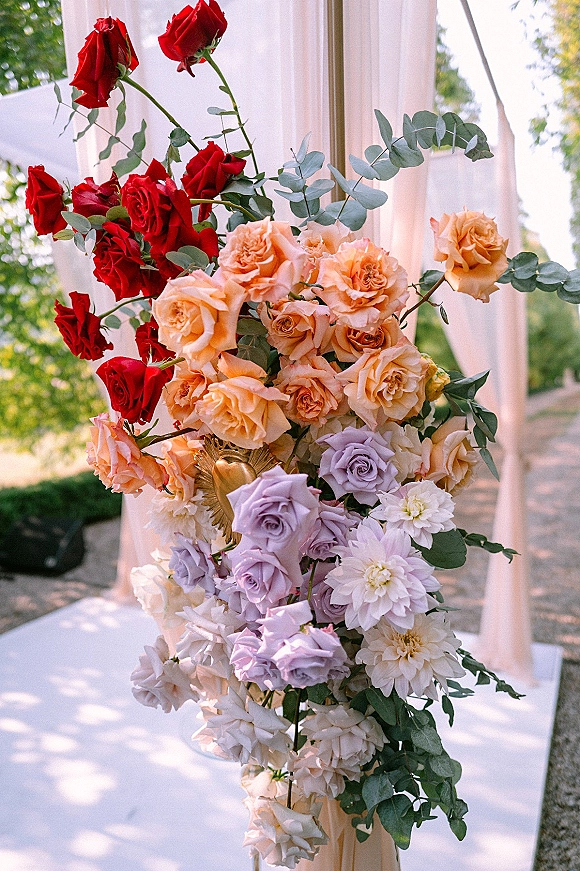 Wedding altar flowers with ceremony arch florals in multicolor roses, eucalyptus, and blush drapery on a white platform in a garden setting