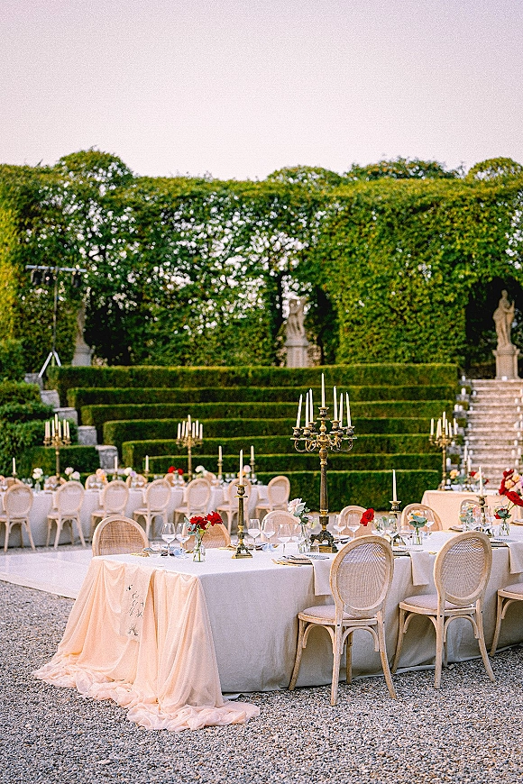 Reception tablescape with outdoor reception tables dressed in white linen, brass candelabras, red roses, and taper candles on a gravel garden patio