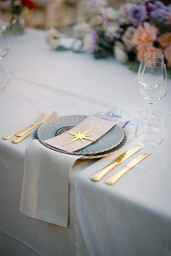 Wedding place setting with gold flatware, blue charger plate, and menu card on a white tablecloth, with blurred floral centerpiece behind