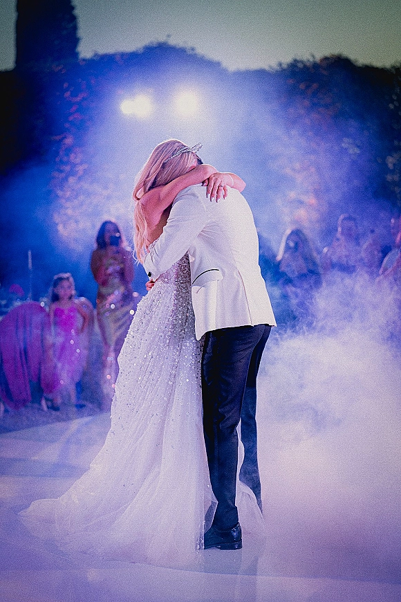 First dance as bride in a wedding gown and tiara slow dances with groom in a white tuxedo jacket amid fog under stage lights outdoors at night