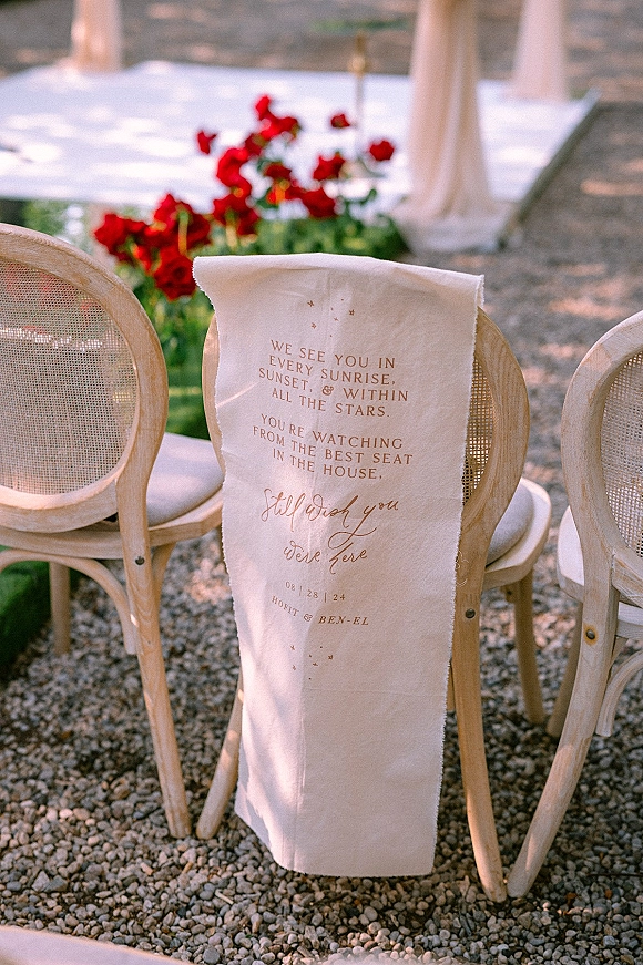 Wedding ceremony chair decor with an aisle chair sign in calligraphy on fabric draped over cane-back chairs by a gravel aisle runner