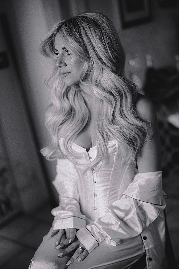 Bridal portrait of a bride in a satin robe and corset top, showing her engagement ring and manicure in a softly lit room