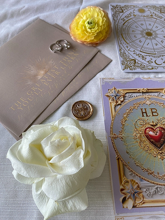 Wedding invitation suite with gold foil lettering, ornate taupe cards, wedding rings, wax seal, and a white rose on linen backdrop