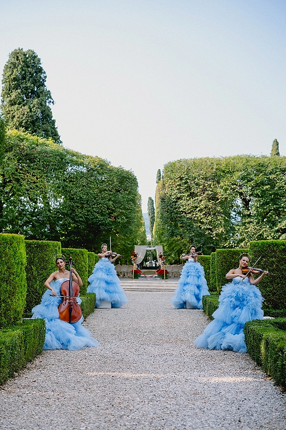 Wedding ceremony musicians in blue tulle gowns play violins and cello along a formal garden aisle with hedges and a draped arch