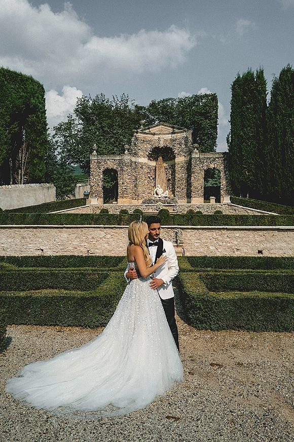 Couple portrait of bride and groom embrace, her strapless gown with long train beside a stone fountain and statue in formal gardens