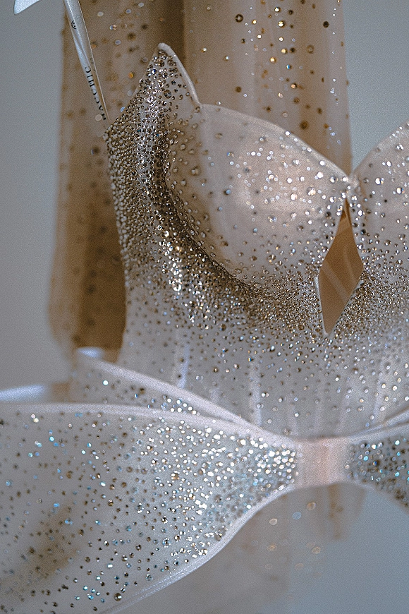 Wedding dress close-up with crystal embellished wedding dress beading, rhinestones, satin bow straps on a hanging gown against a blurred indoor backdrop
