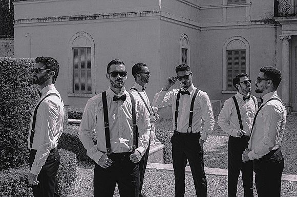 Groomsmen portrait of a stylish lineup in suspenders, standing in a gravel courtyard by a villa with shuttered windows and hedges