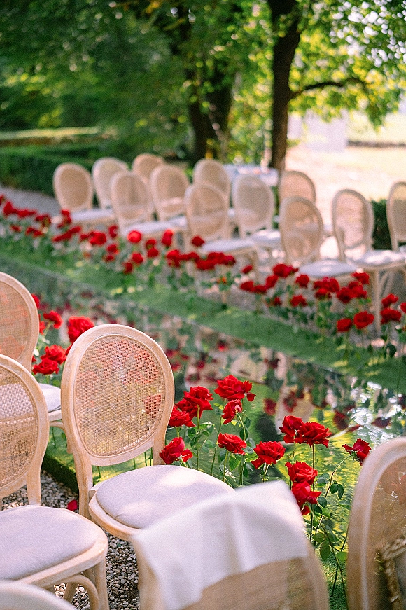 Ceremony aisle decor with red roses lining a mirrored aisle runner, flanked by white cane-back chairs on a sunlit garden lawn