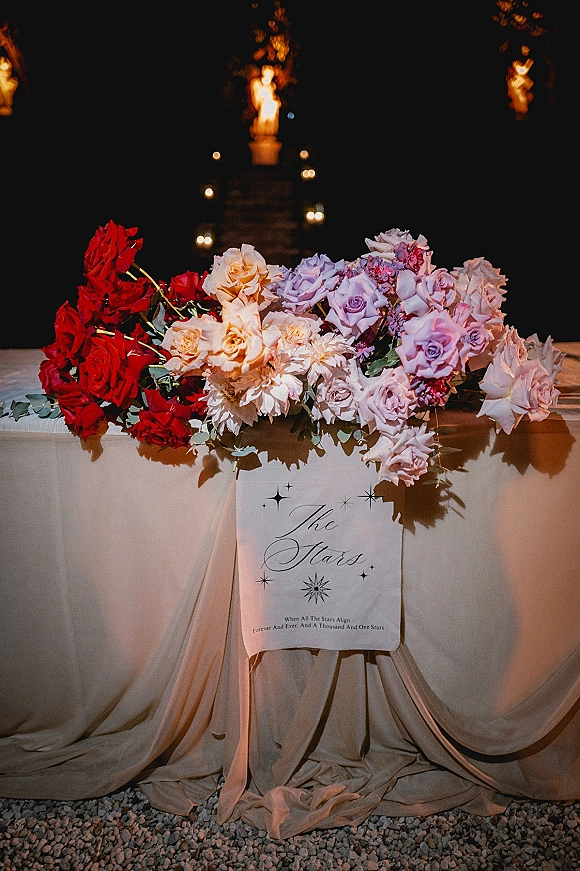 Wedding welcome sign with flowers atop a draped table, framed by red and lavender roses and greenery beside a candlelit walkway at night