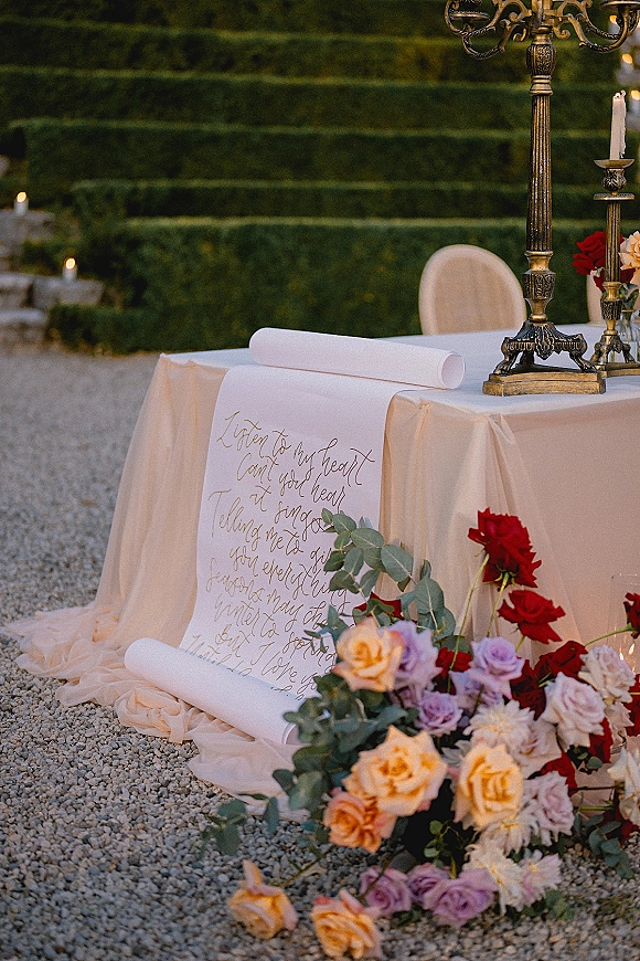Wedding ceremony decor with a wedding calligraphy scroll beside brass candelabra, taper candles, and rose-eucalyptus florals on gravel by hedge steps