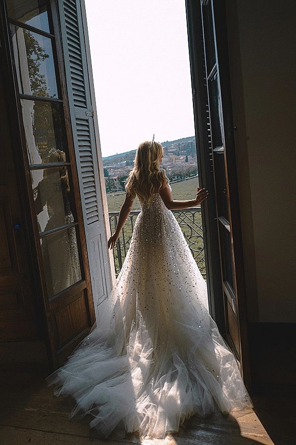 Bridal portrait of a bride at window in a sparkly ball gown with tiara and flowing tulle train, looking out to a hillside balcony view