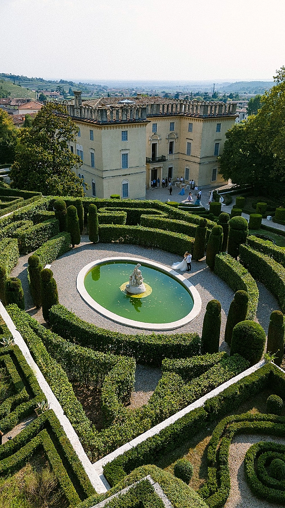 Wedding venue with formal garden hedges and fountain statue, gravel paths leading to a historic villa, with hillside town beyond