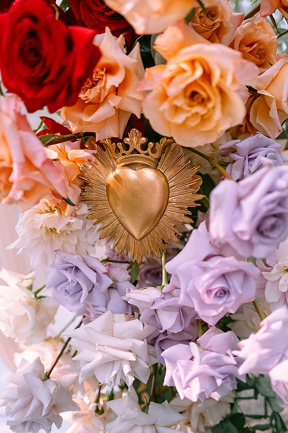 Wedding floral arrangement of colorful wedding flowers with roses, greenery, and a gold sacred heart ornament against softly blurred florals