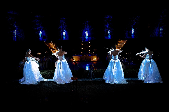 Wedding violinists perform live music in glowing ball gowns with tiaras by a candlelit stone staircase at an outdoor garden reception at night