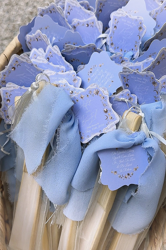 Wedding favor fans with custom wedding hand fans tags, blue ribbon and white tassels, celestial star-and-moon print and gold foil lettering in close-up stack
