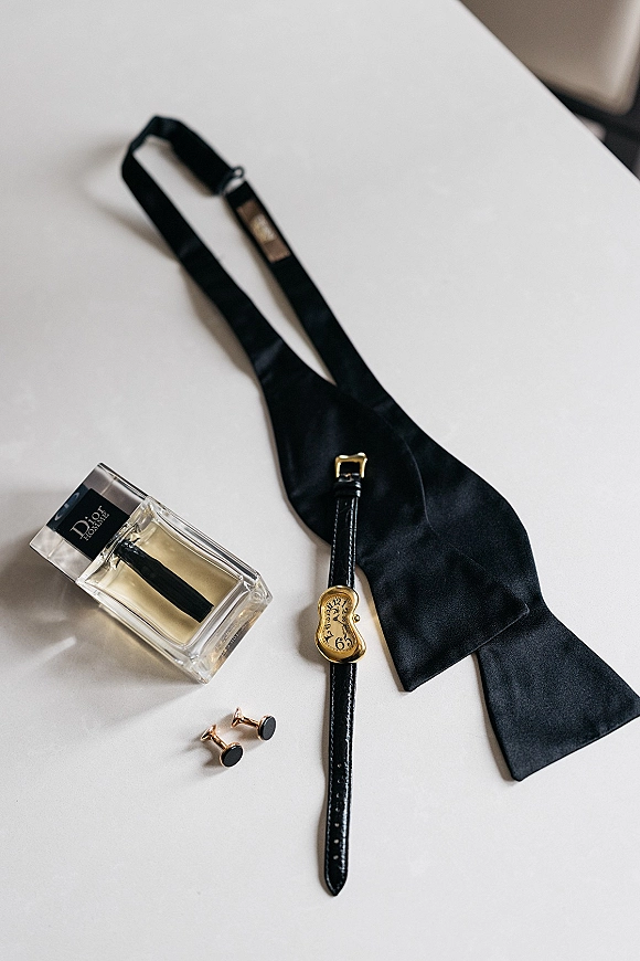 Groom accessories flat lay featuring a black bow tie with wristwatch, cufflinks, and cologne bottle arranged on a white surface