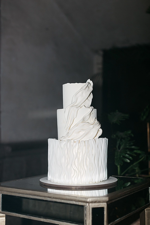 Wedding cake with sculptural textured buttercream ruffles on a mirrored table, three-tier white design against a dark indoor wall with greenery