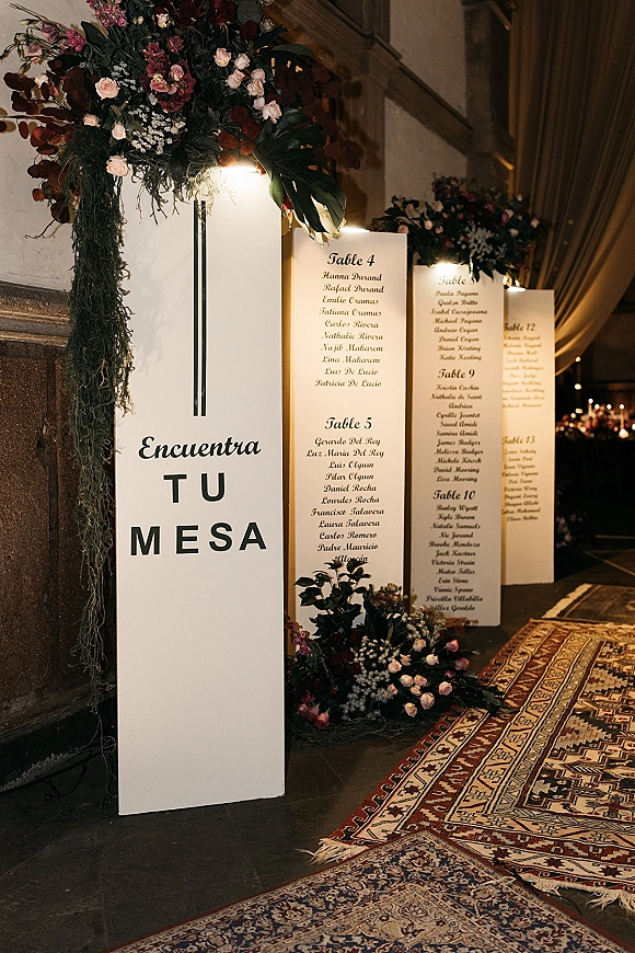 Wedding seating chart on acrylic panels with printed guest cards, framed by rose and greenery floral accents in a draped hallway with rugs
