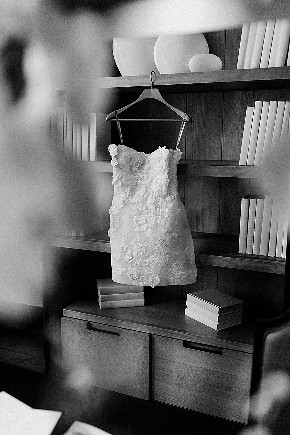 Wedding dress, lace wedding dress hanging on a hanger with floral appliqué beside books, set against a wood bookshelf and drawers