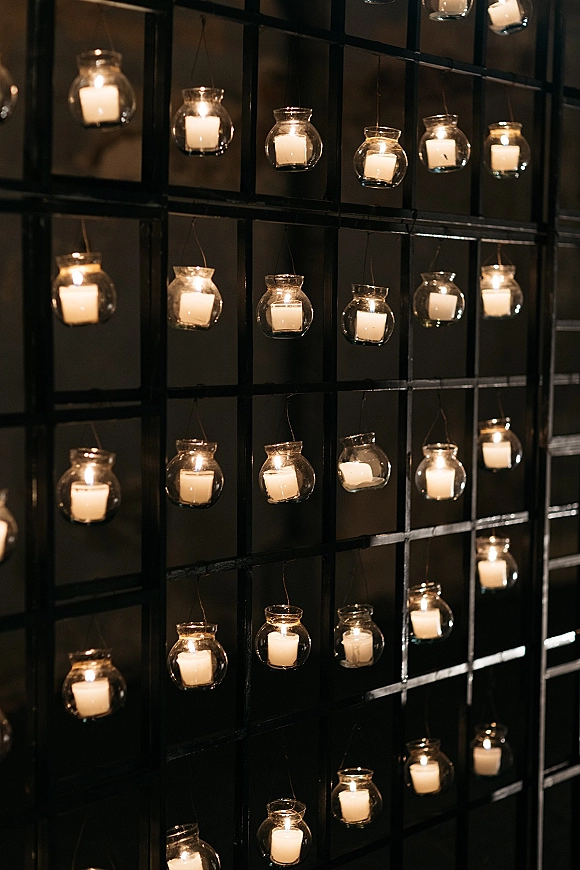 Hanging candle decor with hanging glass candle holders in glass votive holders, suspended on wire against a black grid wall in a dark interior