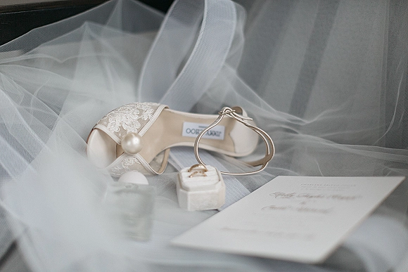 Wedding shoes lace wedding heels with pearl ankle straps beside an engagement ring, ring box, and invitation on a soft gray flat lay