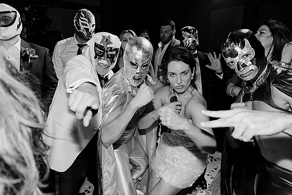 Wedding reception dancing as bride and groom in luchador masks and capes celebrate with guests amid confetti on an indoor dance floor