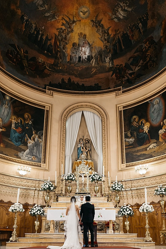 Wedding ceremony with bride and groom from behind at a church wedding altar, priest officiating beneath murals, chandeliers, and candles