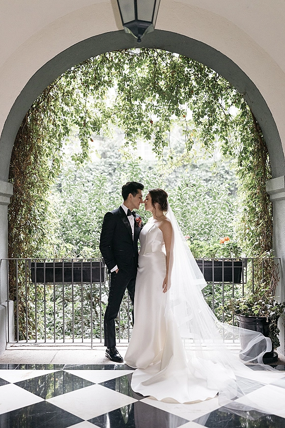 Couple portrait of bride and groom portrait touching foreheads under an ivy-covered archway, her cathedral veil flowing over black-and-white tile floor