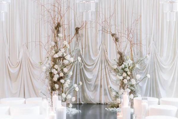 Ceremony setup with a modern wedding altar framed by curly willow branches, set before draped white curtains on a polished concrete floor.