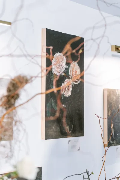 Floral painting of roses, a rose oil painting illuminated by a brass picture light on a white wall amid additional artworks.