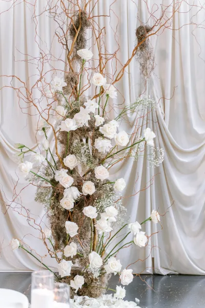Ceremony floral installation of wedding altar flowers with white roses arranged before draped white fabric on a polished gray floor.