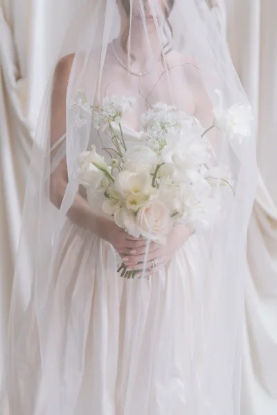 Bridal bouquet, a white rose bouquet gently tucked beneath a sheer veil, set against a soft cream drapery backdrop.