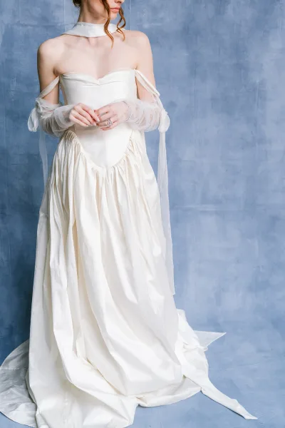 Wedding dress close-up of an off-the-shoulder wedding dress with sheer gloves against a blue studio backdrop.