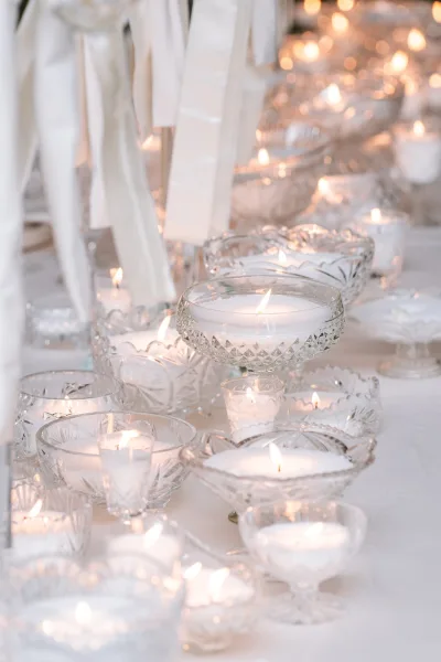 Candle centerpiece featuring crystal candle holders clustered along the table, trimmed with white ribbons, casting a soft, romantic glow.