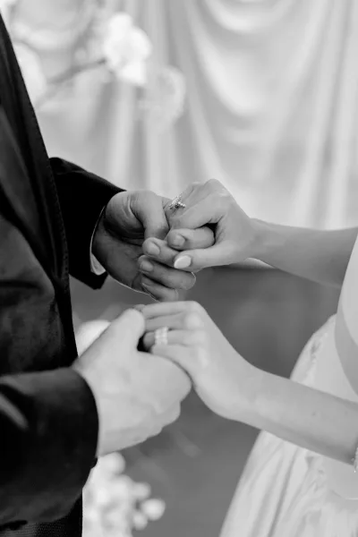Ceremony moment with the bride holding groom's hand, a sparkling engagement ring in a close-up against draped fabric and florals.